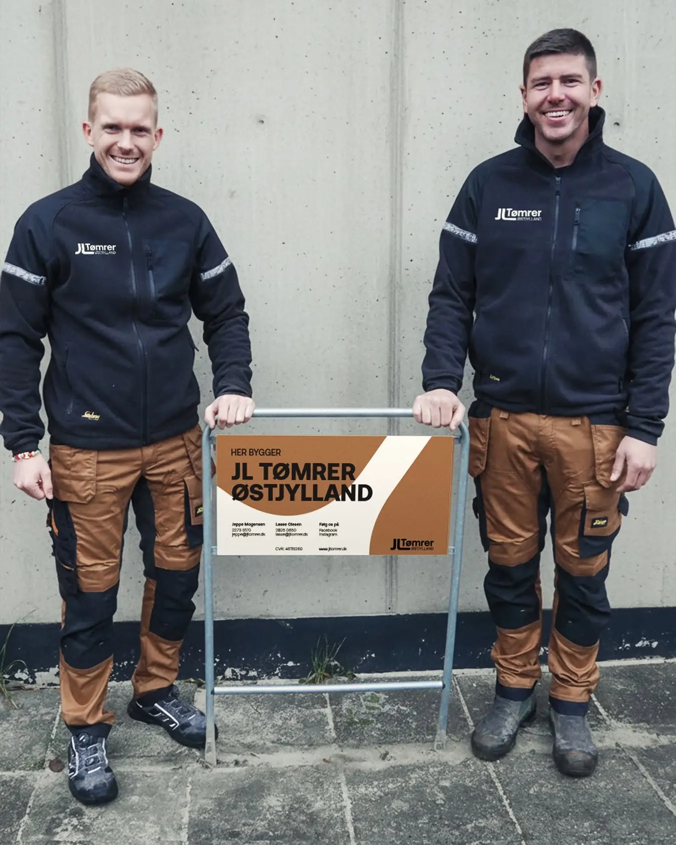 Lasse Olesen og Jeppe Mogensen i arbejdstøj ved deres byggepladsskilt foran en betonvæg.
